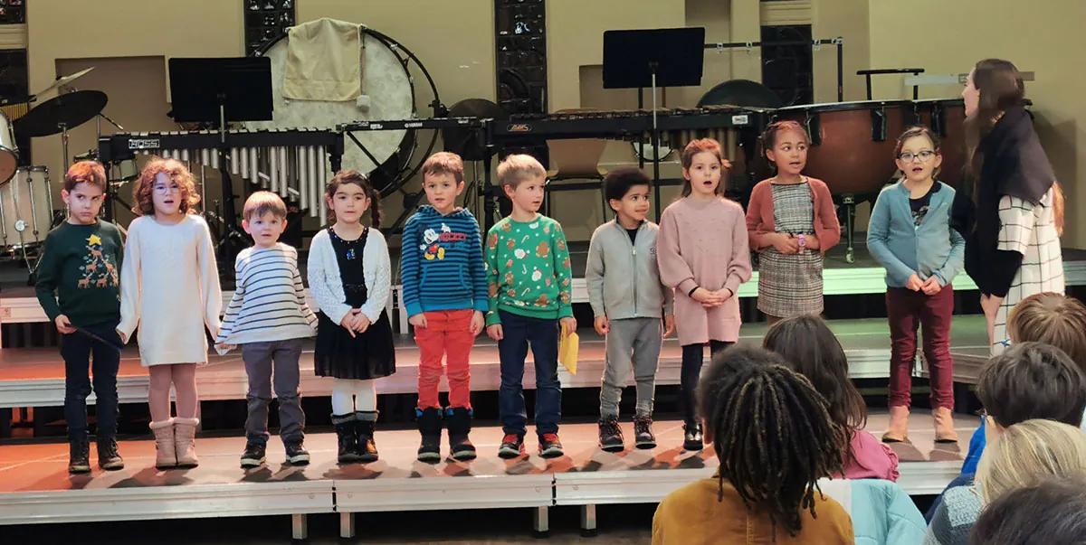 Un groupe de jeunes enfants chantant sur scène avec une intervenante — une audition d’éveil ou d’initiation musicale, printemps 2025