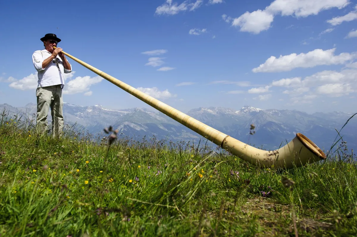 Musicien jouant d'un long cor des Alpes en bois dans un paysage montagneux suisse.